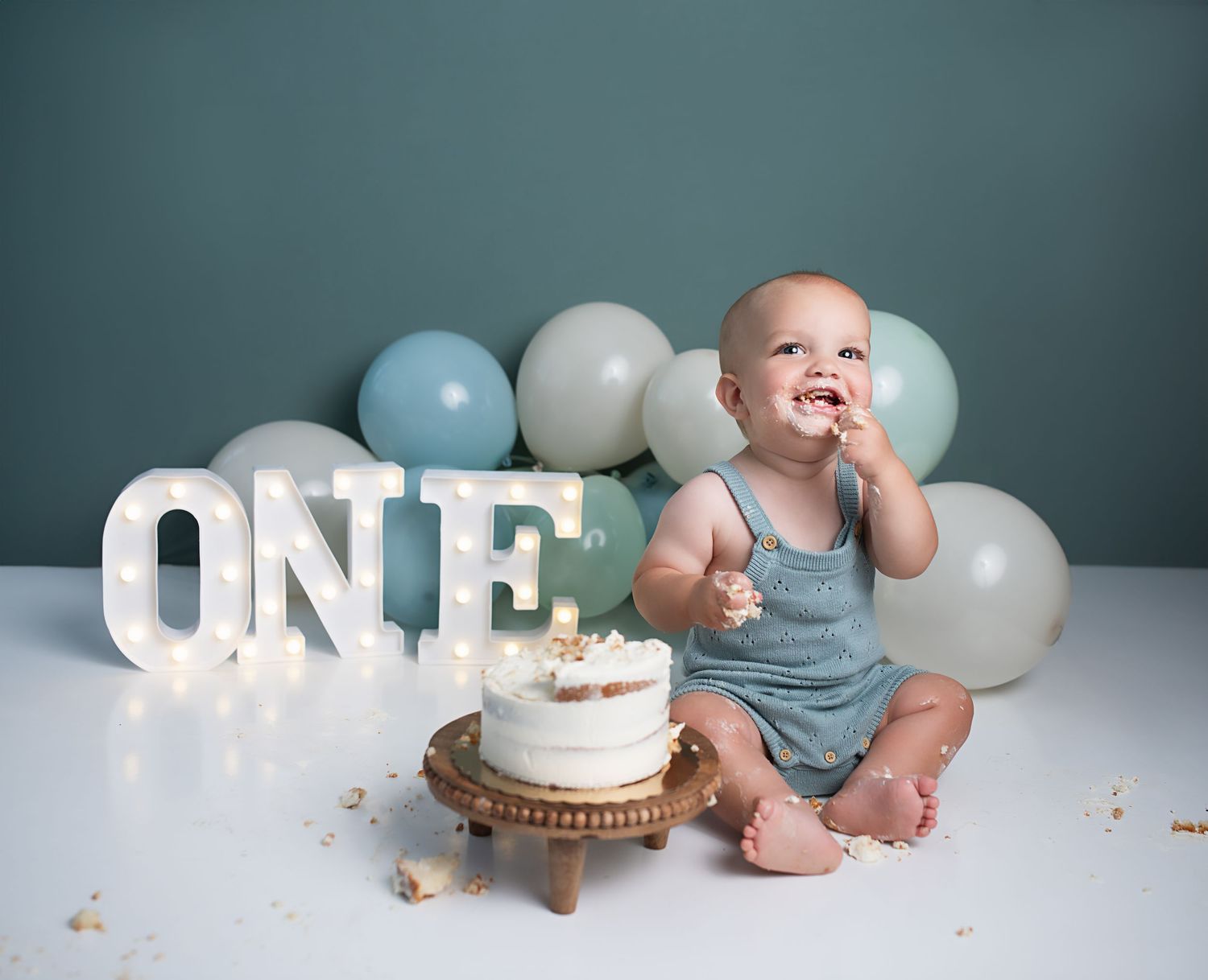 Happy baby boy laughs while eating cake at first birthday cake smash session with blue balloons backdrop.