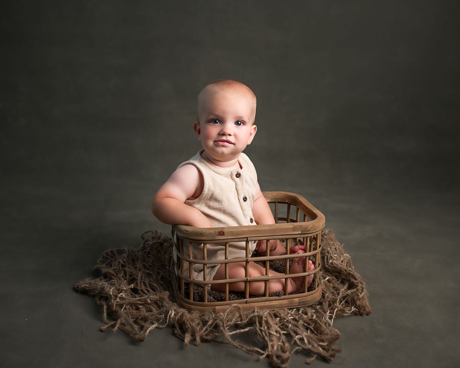 Cute baby in cream romper sitting in wicker basket with colorful eggs on green backdrop.
