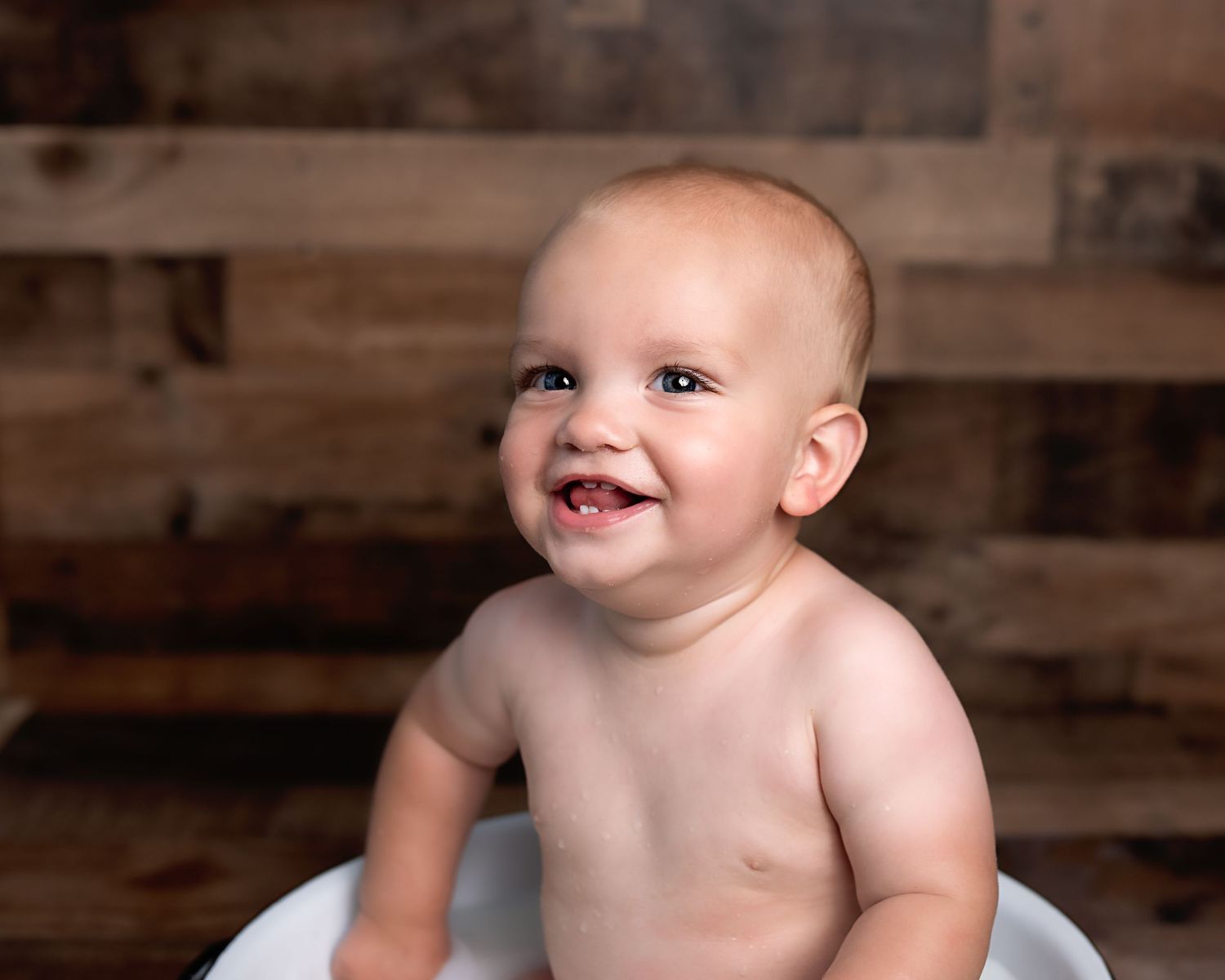 Smiling baby boy with two teeth sits in white tub against rustic wood backdrop in studio portrait.