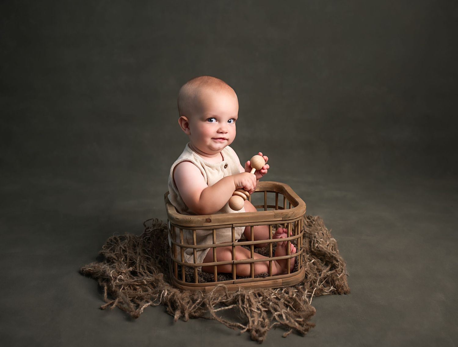 Smiling baby sitting in rustic wicker basket holding toy giraffe on olive background.