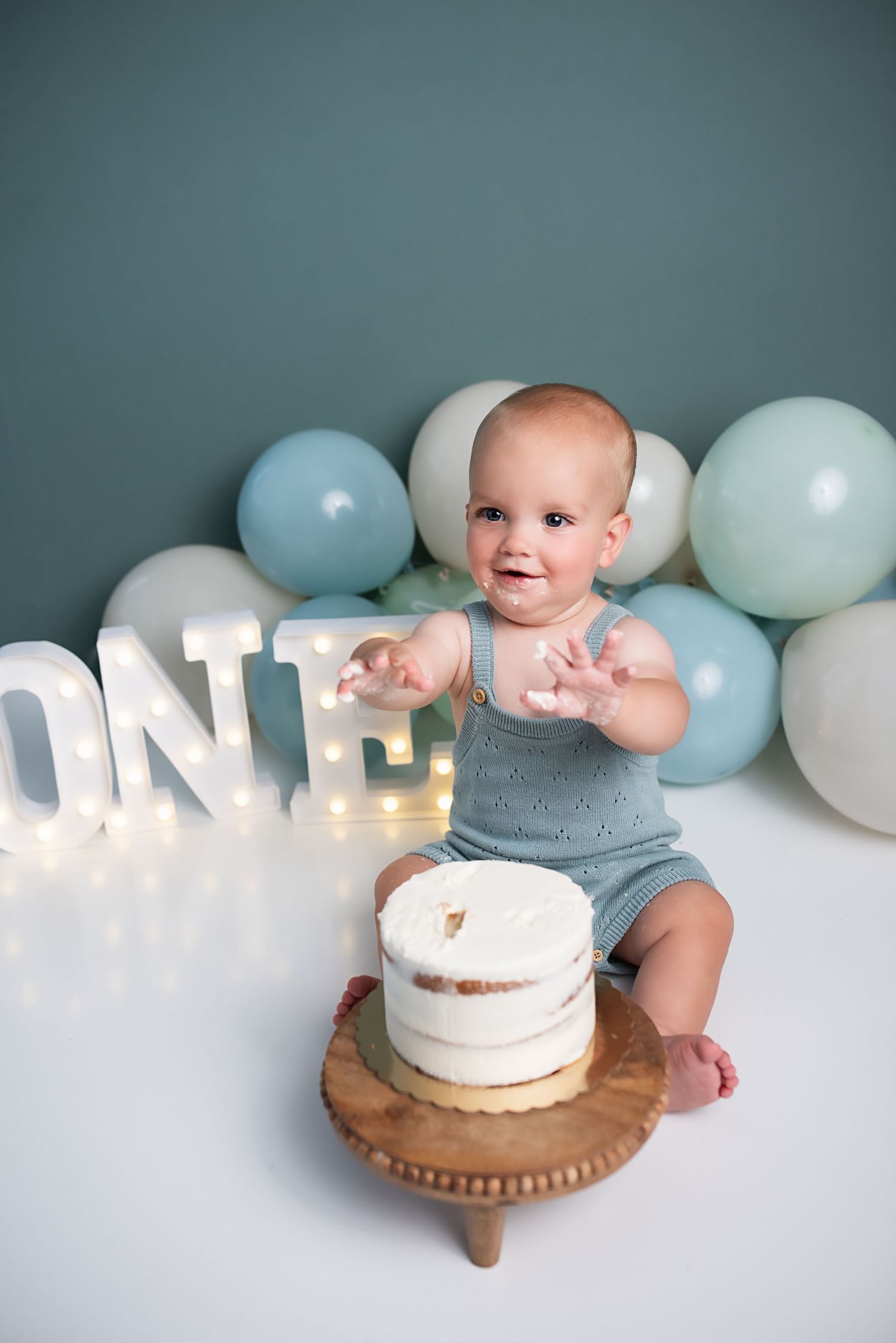 Smiling baby boy claps frosting-covered hands at his first birthday cake smash session with ONE sign.