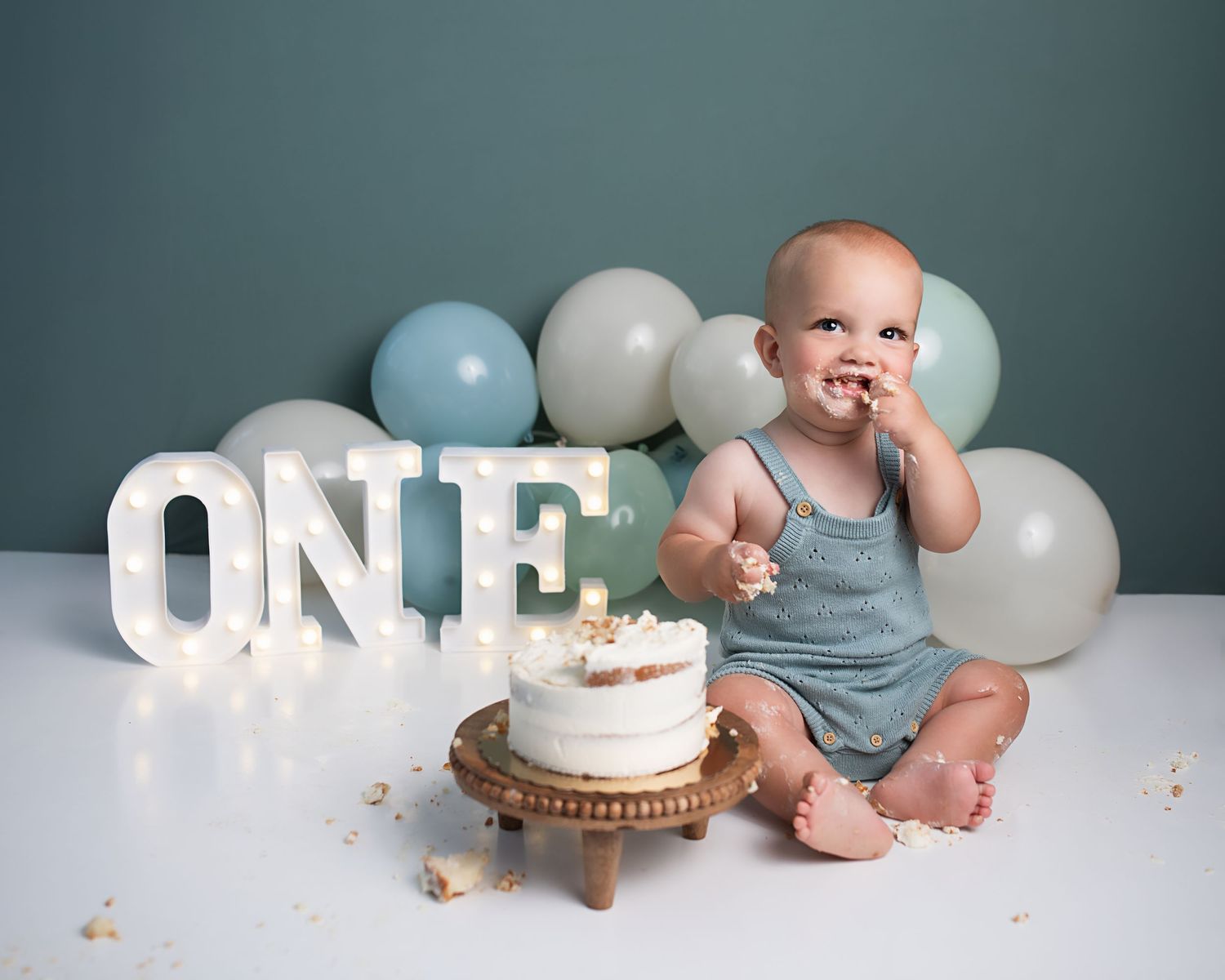 Baby boy in gray romper eats birthday cake at first birthday cake smash session with lit ONE sign.