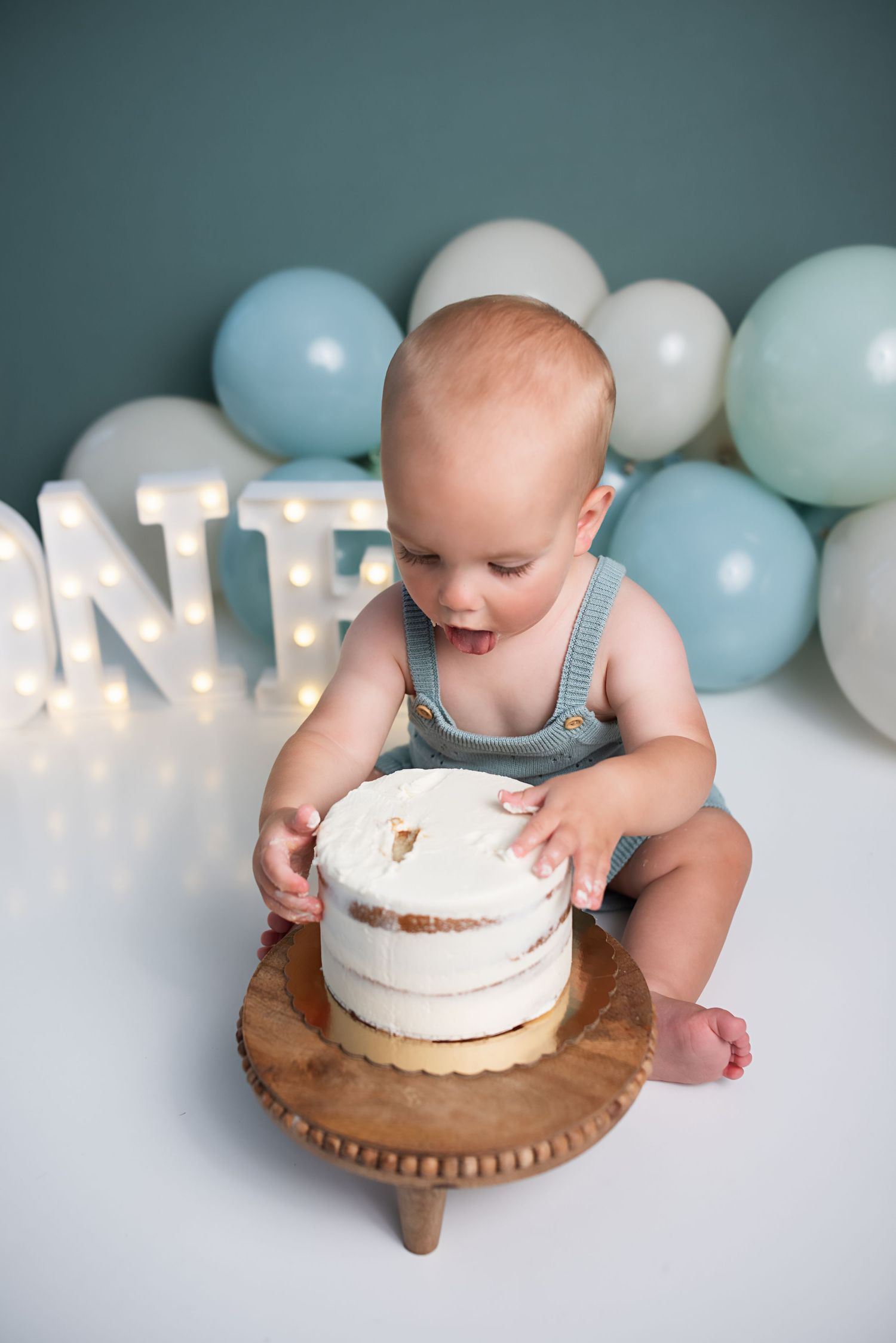Baby boy in gray romper touches his first birthday cake at a cake smash photo session with blue balloons.