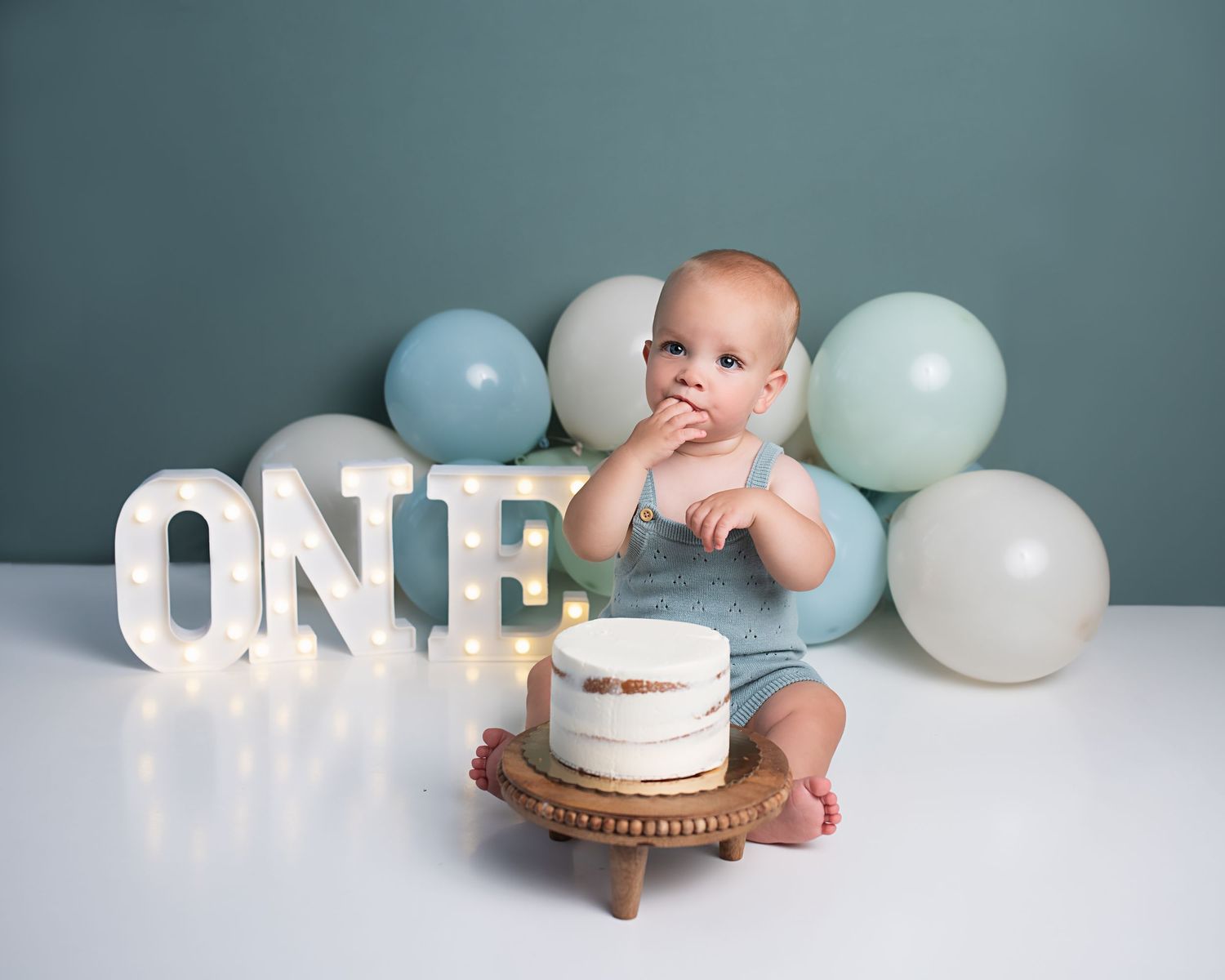 Baby in blue romper sitting before first birthday cake with ONE sign and pastel balloons.