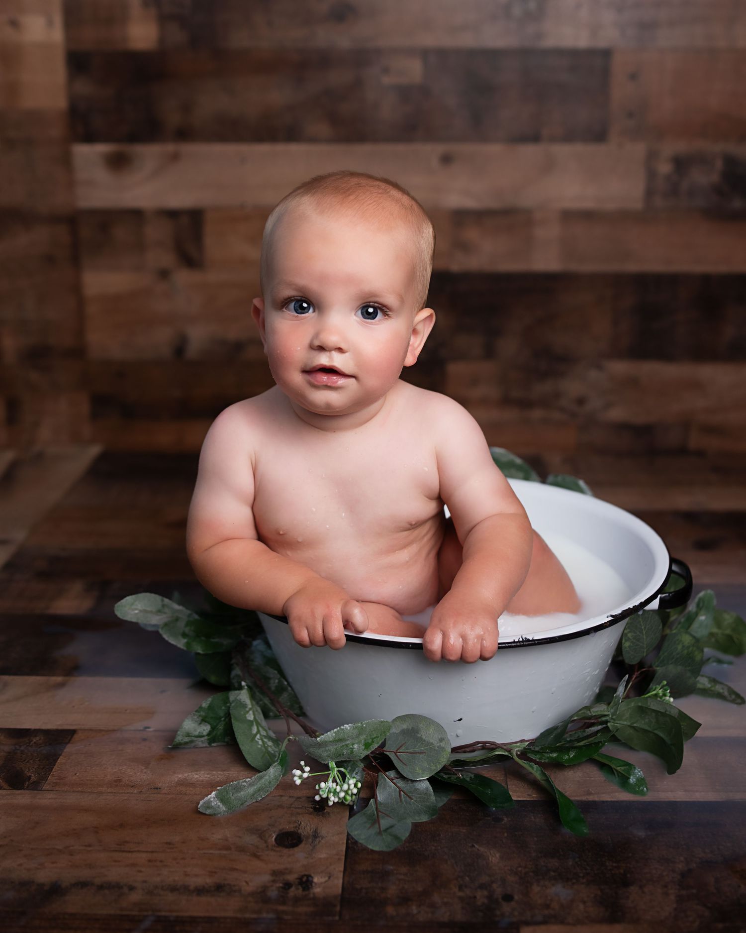Cute baby boy sits in white enamel tub surrounded by greenery against rustic wood plank background.