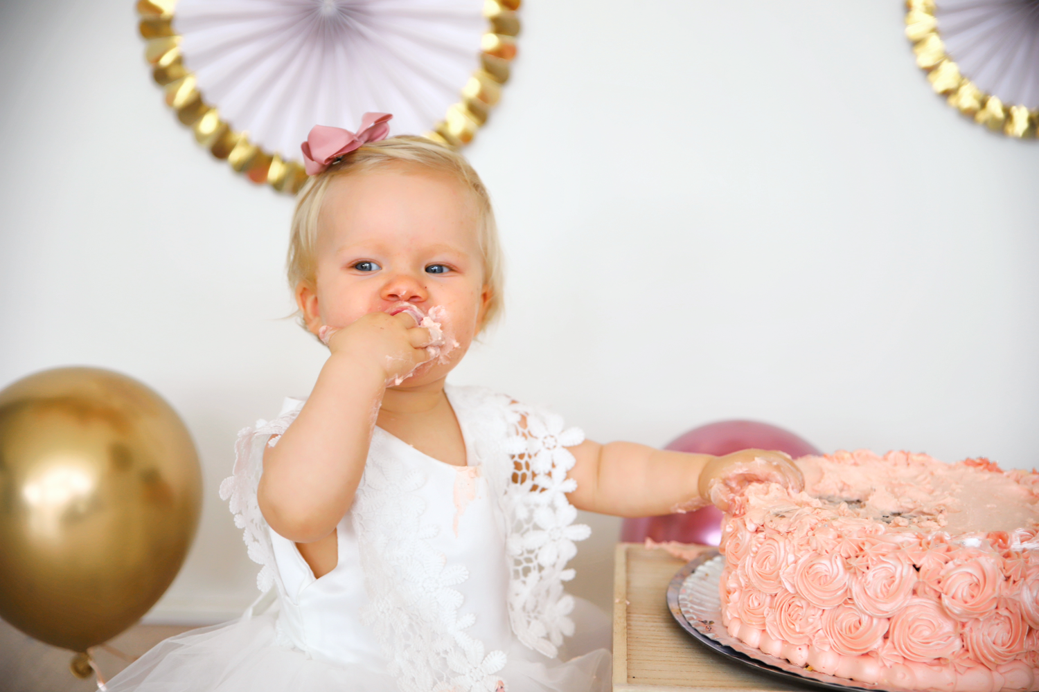 Small child in white dress tastes frosting from pink birthday cake during celebration.