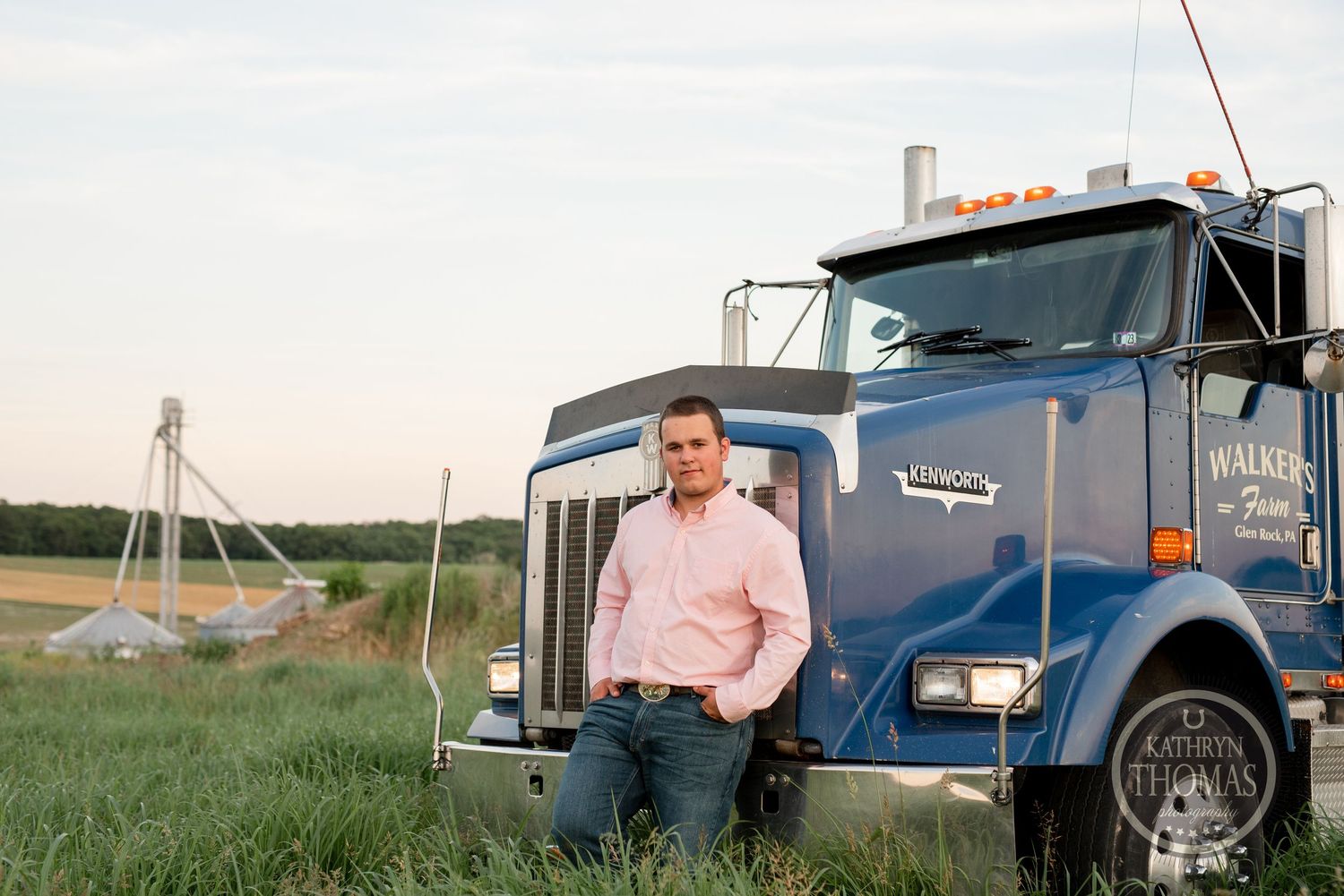 Driver poses on blue Kenworth truck with Walker's Farm branding at dusk.