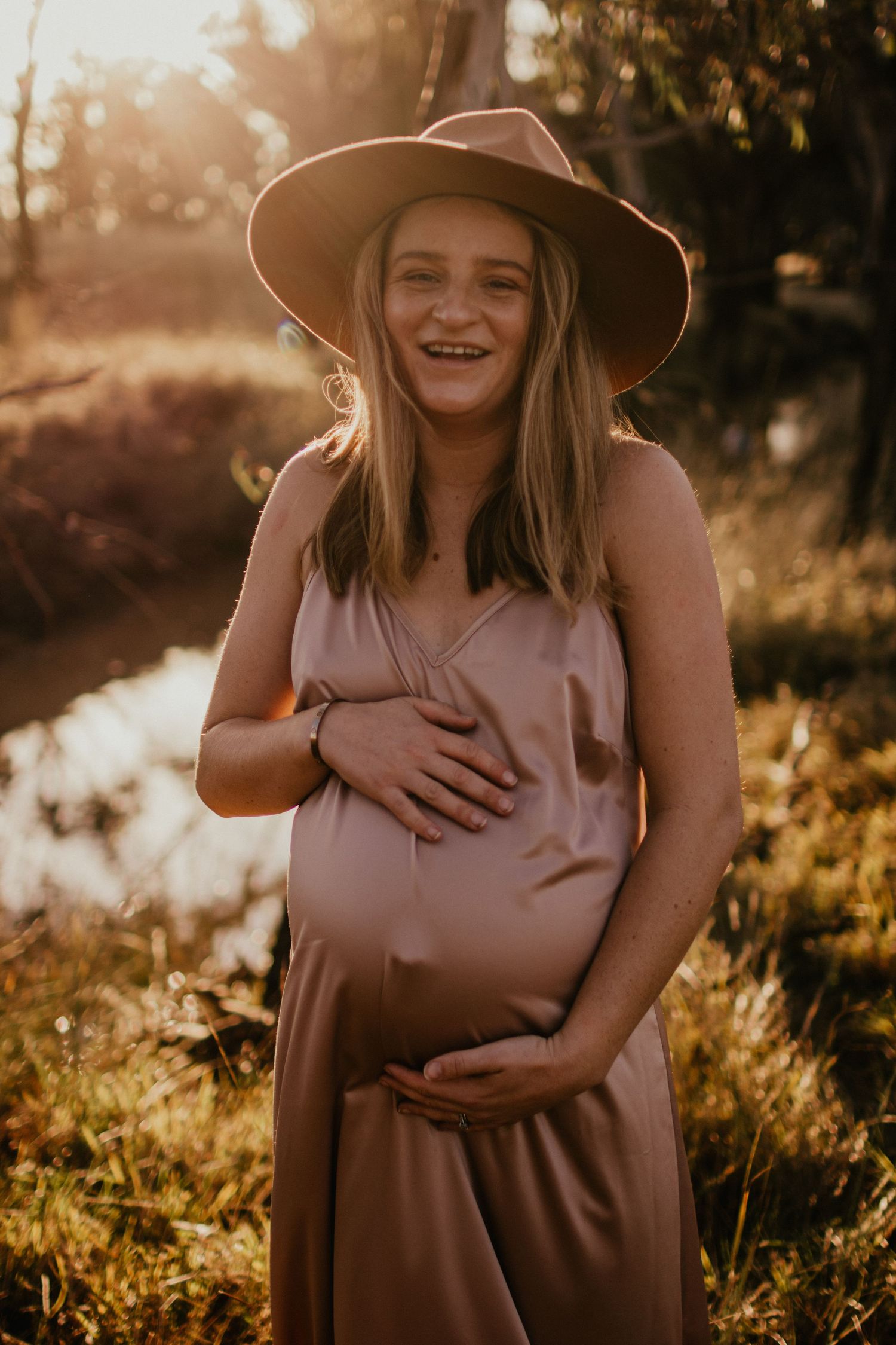 Pregnant woman in a rural setting in a bronze satin dress at golden hour, holding her pregnant belly.