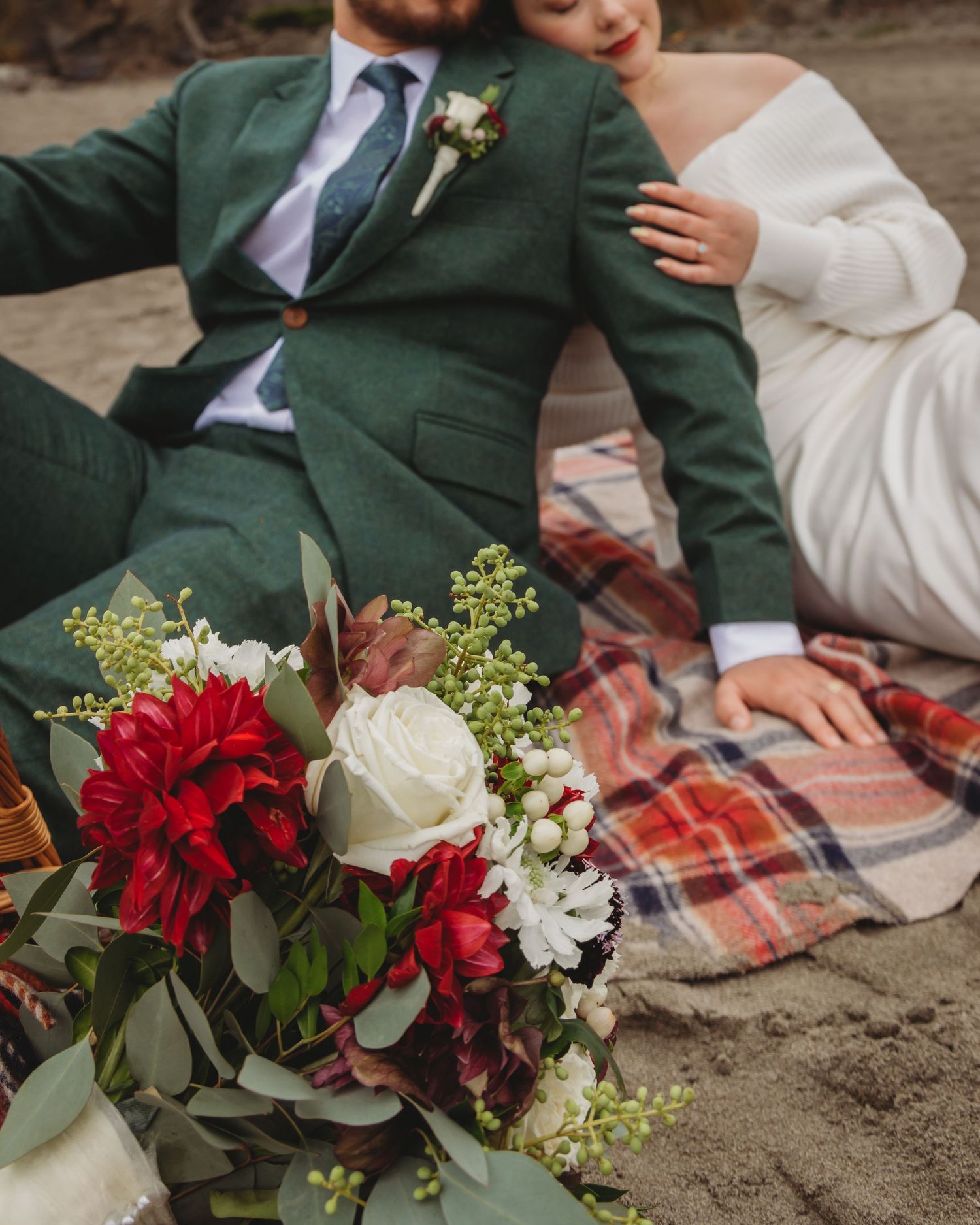 A couple sits on a plaid blanket at the beach with red and white wedding bouquet flowers and greenery.