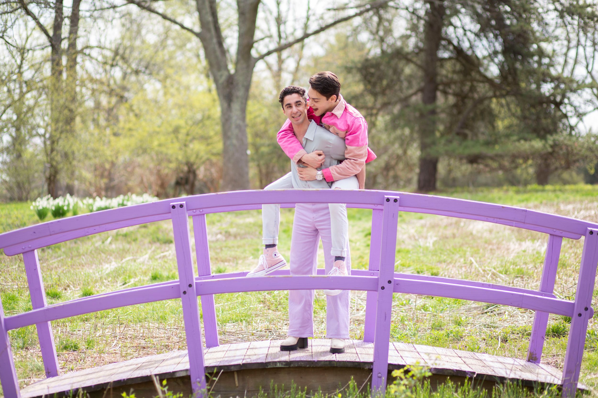 Two people in pink embrace on a purple arched garden bridge surrounded by lush green trees.