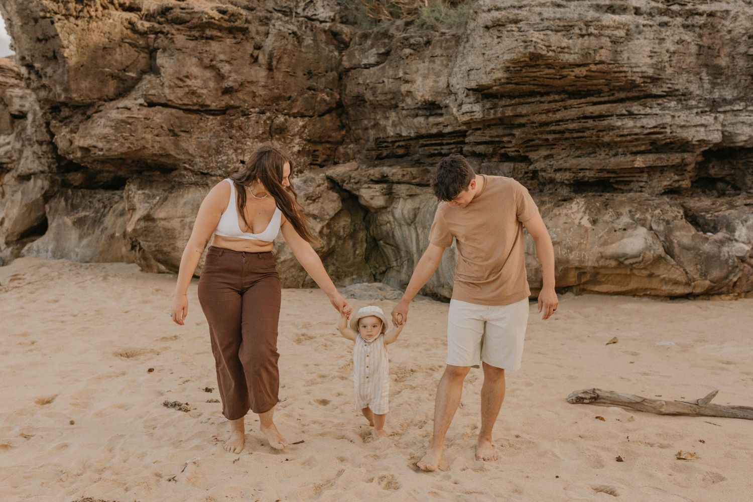 A family walking together on a sandy beach near rocky cliffs while holding their toddler's hands.