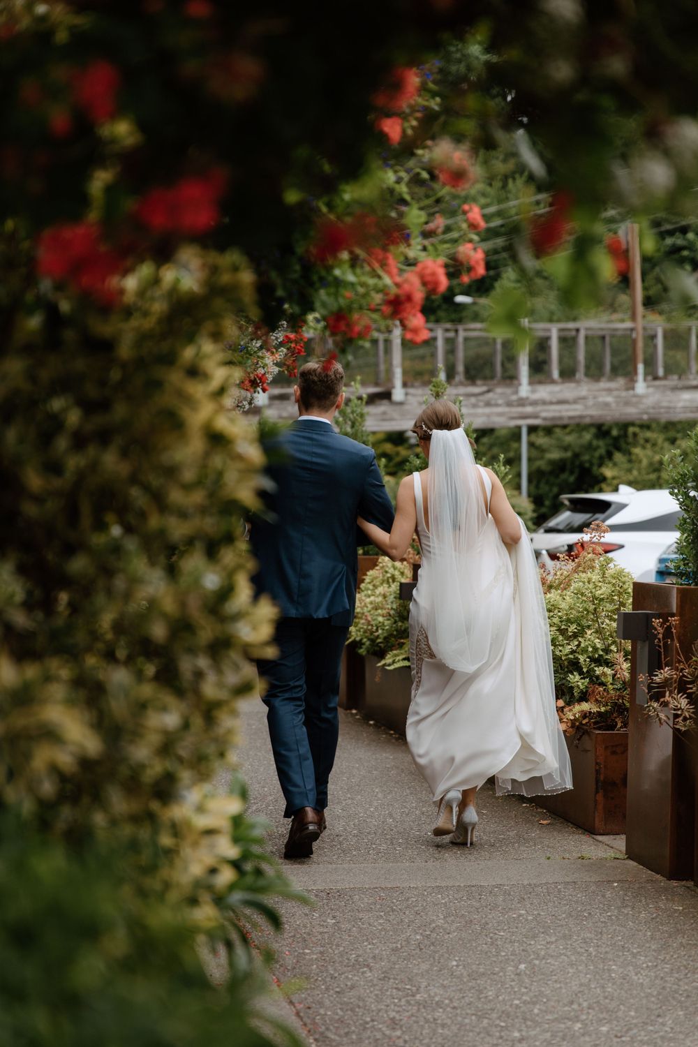 Newlywed couple walking along a garden path under blooming red flowers on their wedding day.