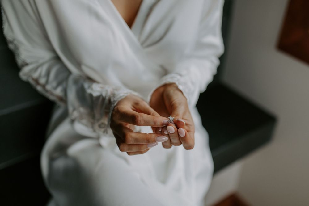 Hands holding wedding rings against white silk robe in soft natural light.