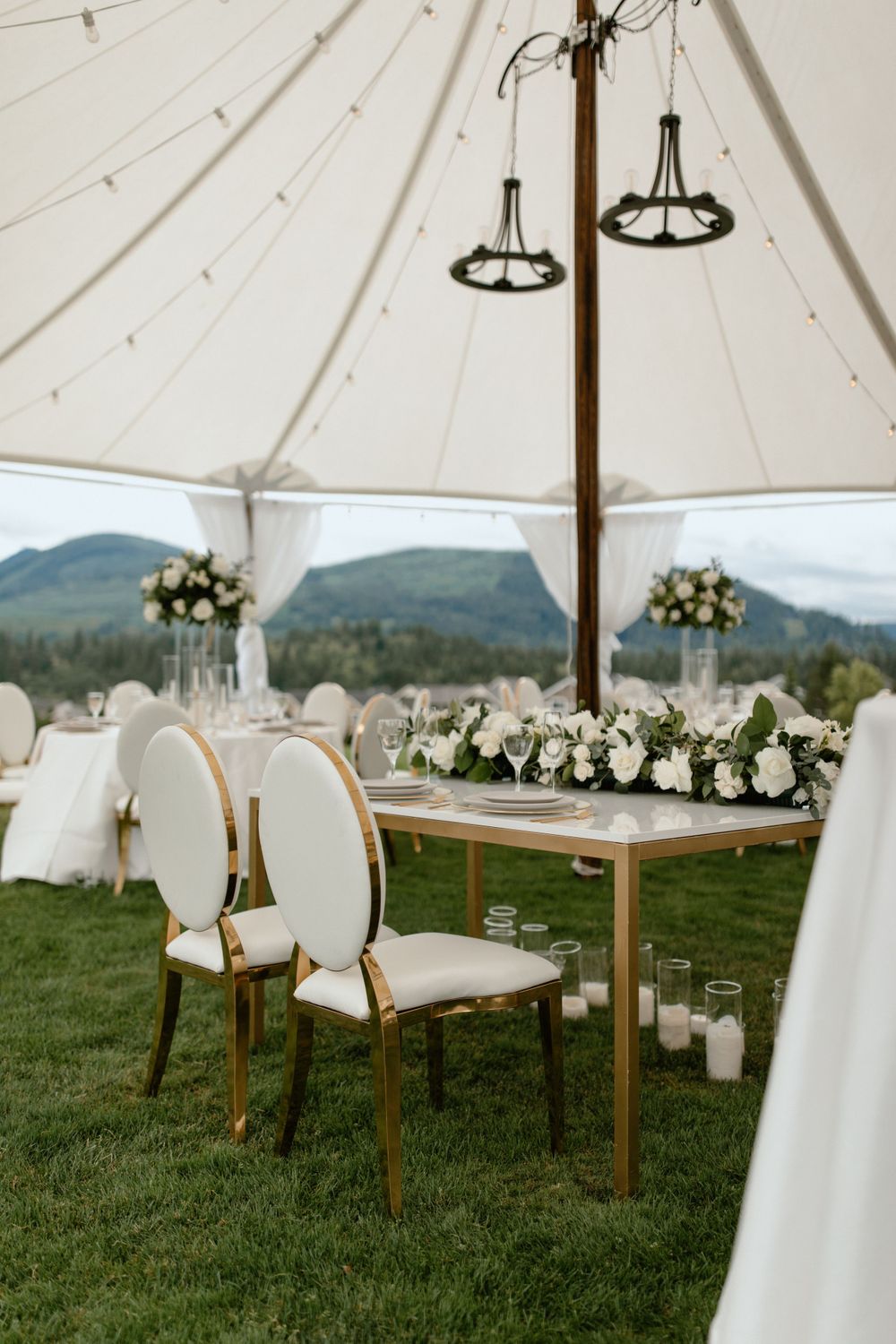 Elegant wedding reception tent setup with white decor and floral arrangements against mountain backdrop.