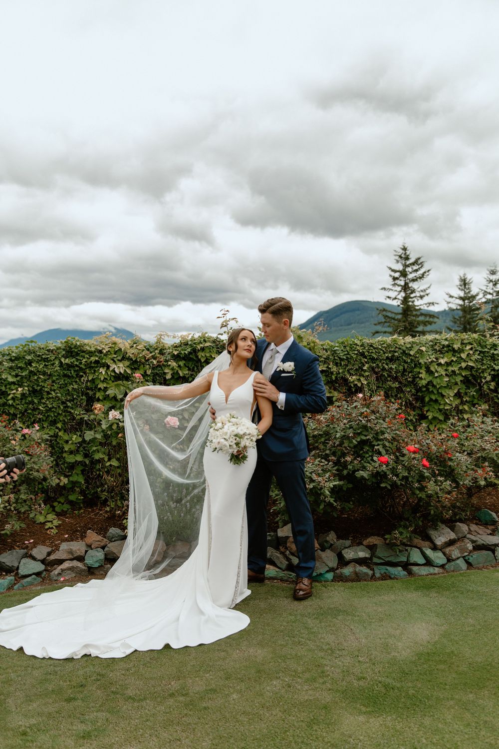 Bride in flowing white gown with long veil poses with groom on scenic garden terrace.