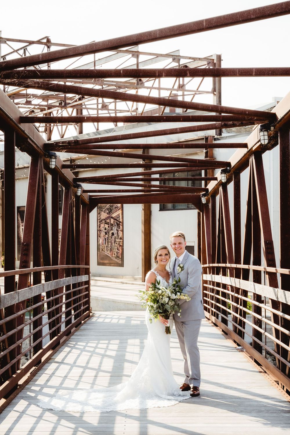 A couple poses for photos on a rustic metal bridge in wedding attire.