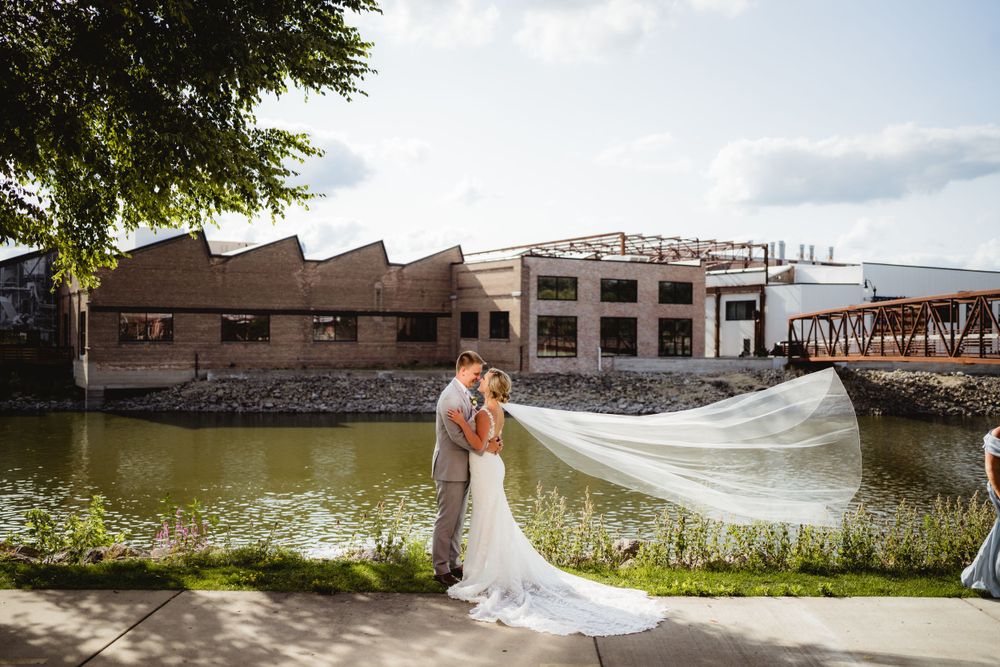A wedding couple poses by a waterfront with their dress veil flowing in the breeze.