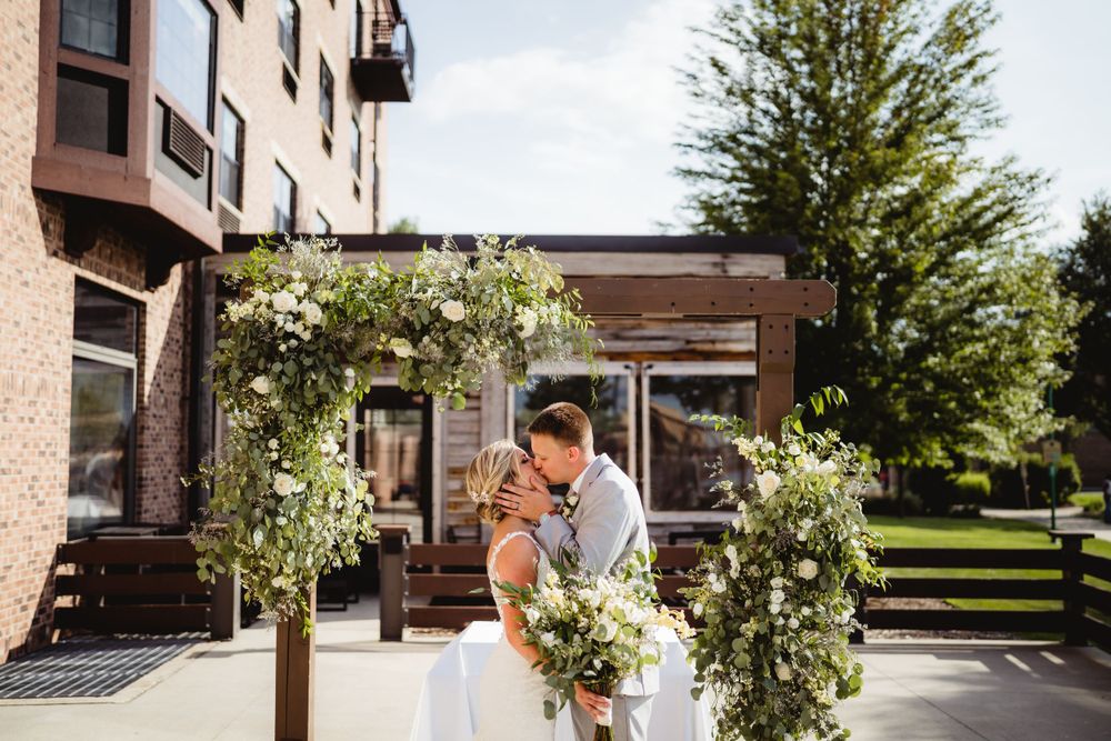 A wedding ceremony takes place under a wooden pergola decorated with white floral arrangements and greenery.