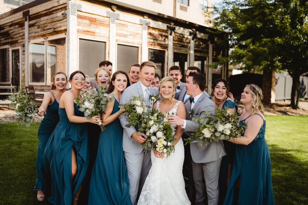 Wedding party in teal dresses and gray suits poses together with bouquets outdoors.