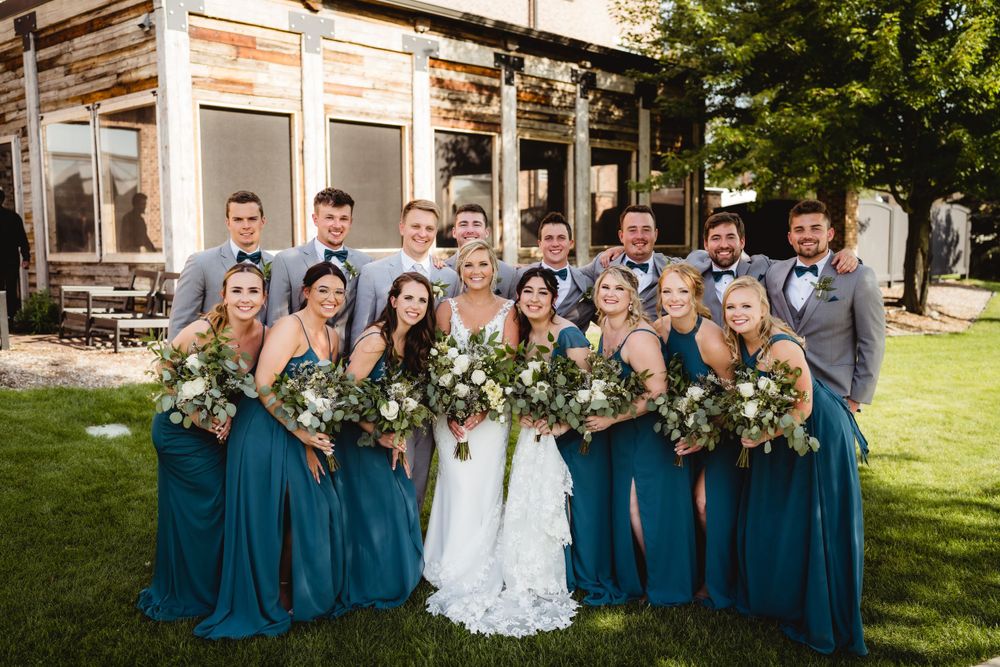 Wedding party in teal blue dresses and gray suits poses in front of rustic wooden building.