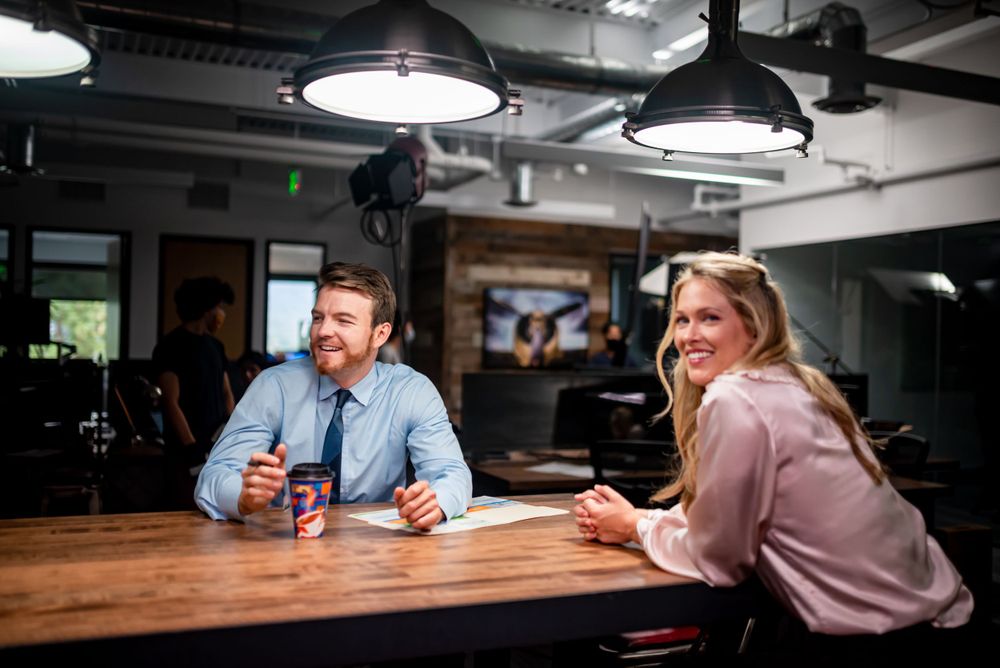 Two people sit at a wooden table in an industrial-style space with pendant lighting.