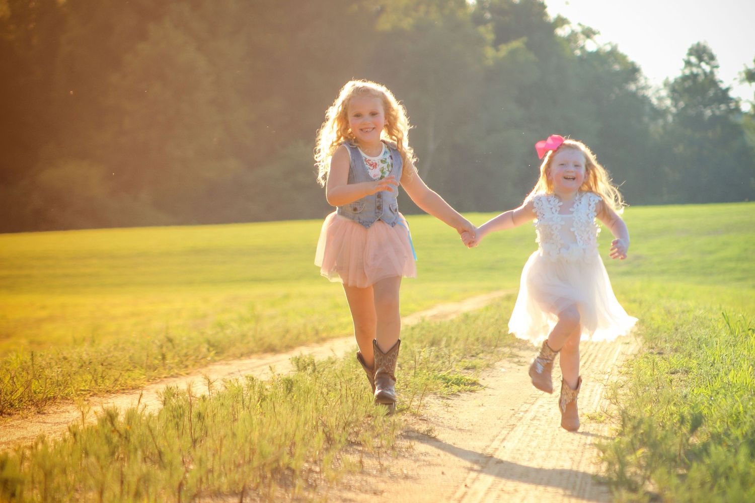 Two children holding hands while joyfully running on a sunlit dirt path through a grassy field.