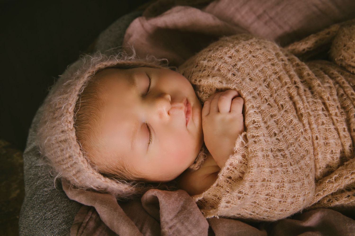 Sleeping baby wearing knit bonnet and wrap in moody lighting.