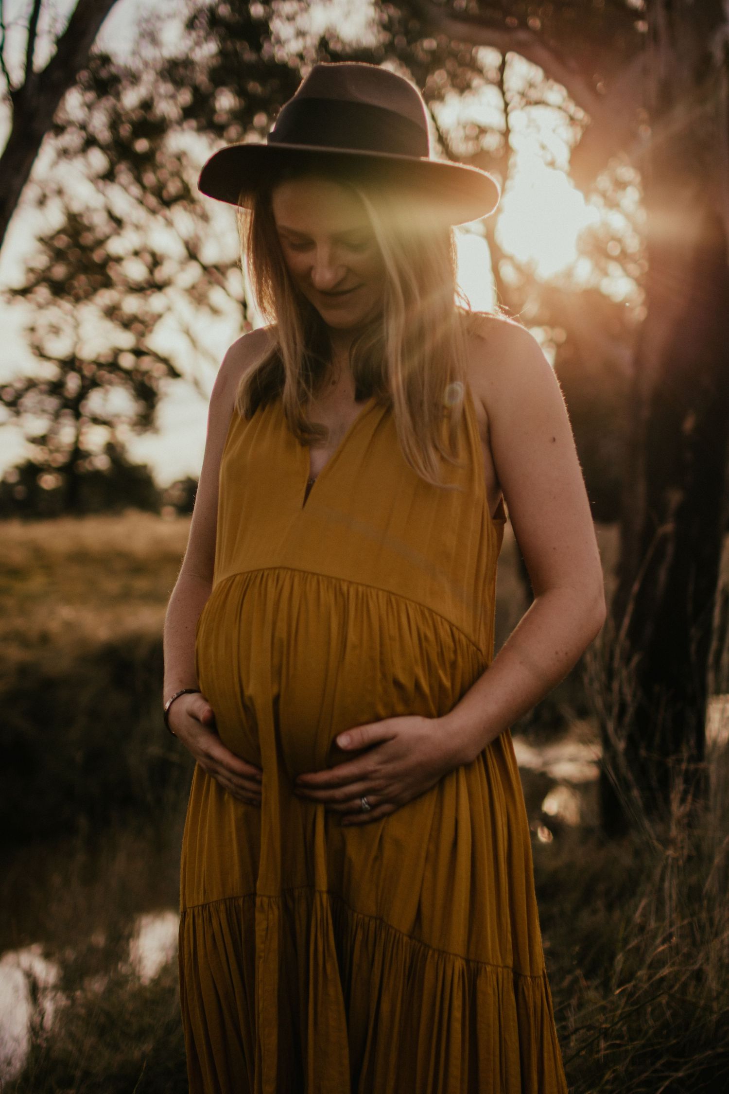 Mum in a golden long dress standing in a paddock with a creek in the background holding her baby belly at golden hour.
