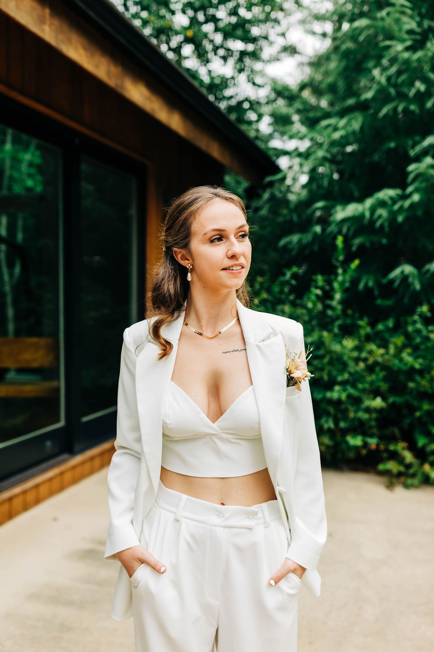 Modern bridal outfit featuring white cropped blazer and pants with floral boutonniere against green backdrop.