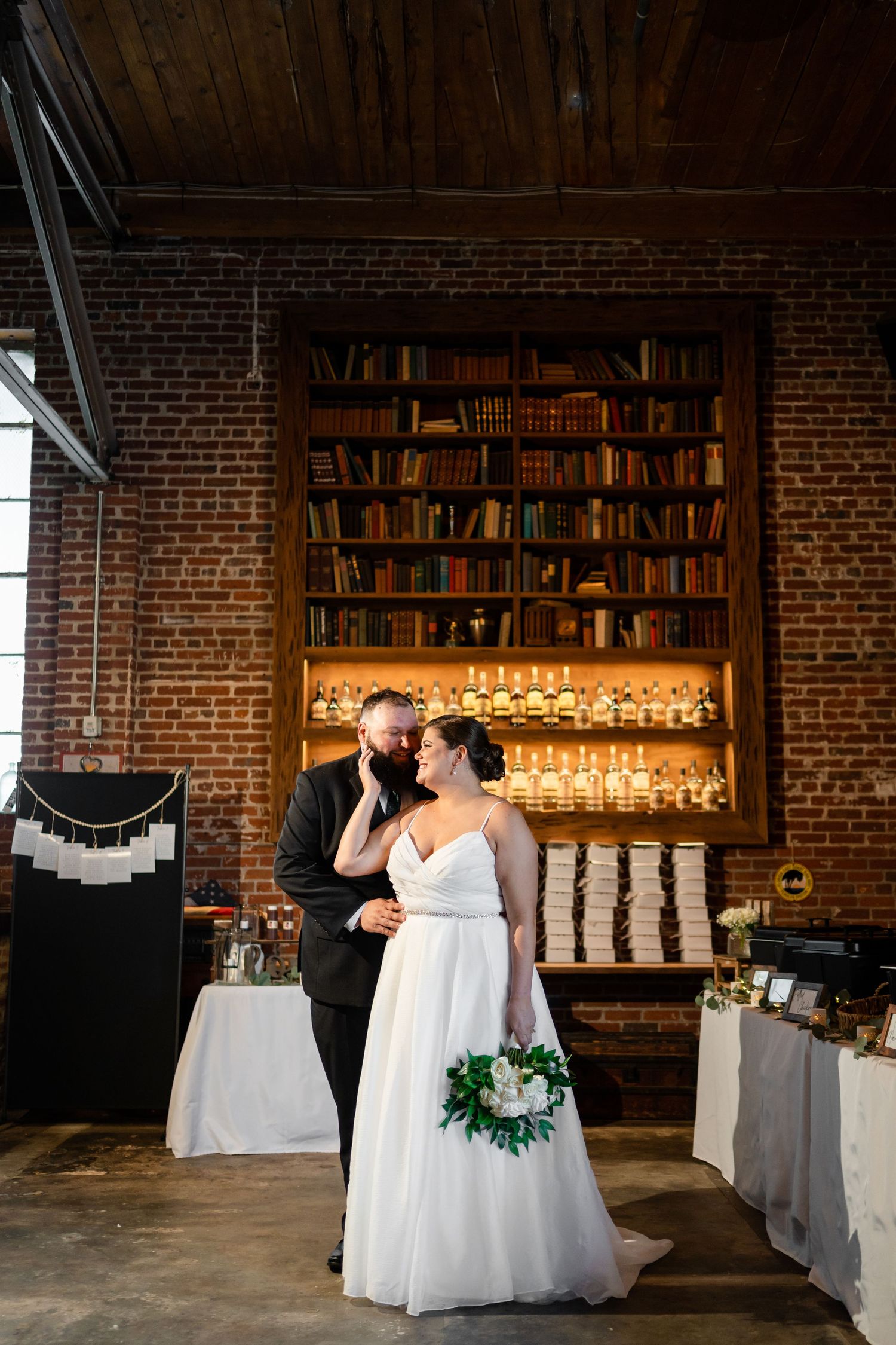 Newlyweds pose in rustic venue with exposed brick wall and library backdrop.