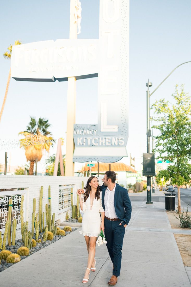 Couple walking together on a palm-lined sidewalk in front of a modern restaurant exterior.