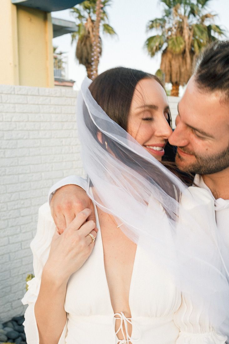 Intimate moment between a couple with a flowing veil against a white brick wall.