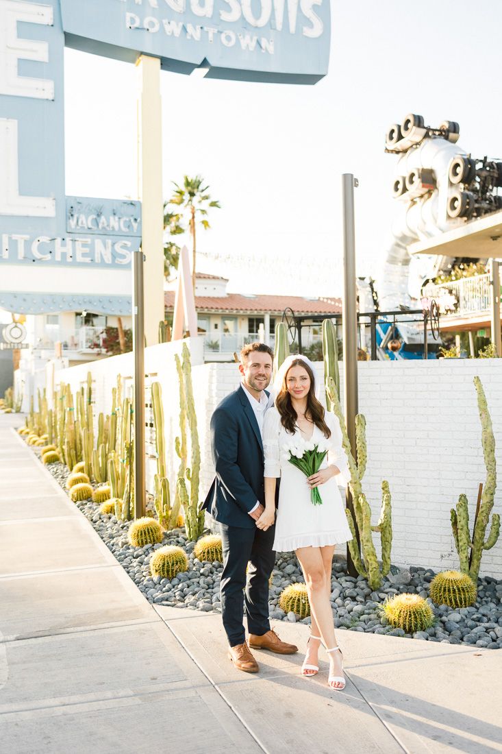 Couple poses together on sidewalk with desert landscaping and palm trees in background.