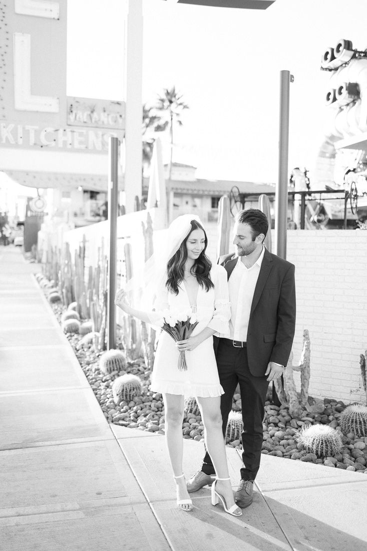A black and white portrait of a couple in formal wear standing on a desert-landscaped sidewalk.