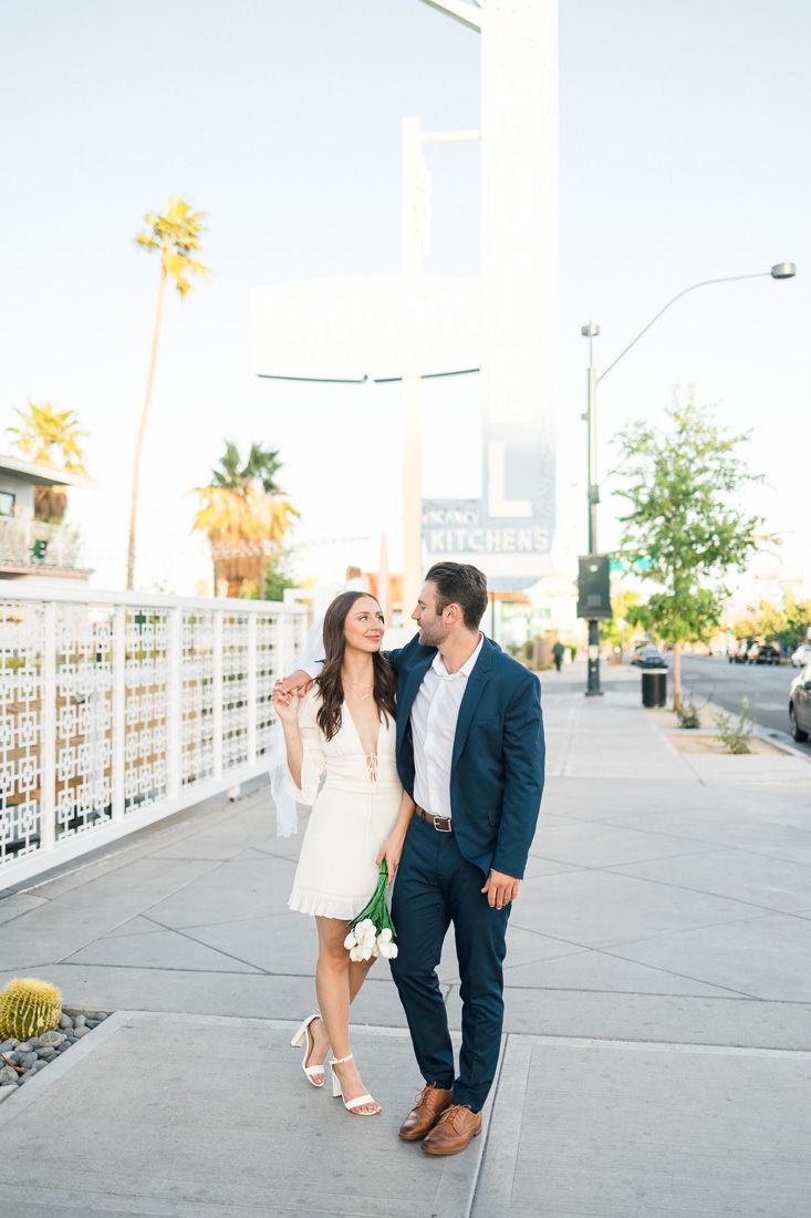 A couple in formal attire walking together on a palm-lined street in a downtown area.
