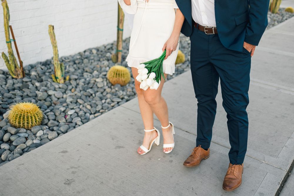 Close-up of formal shoes and attire with white tulips against a modern desert landscape.