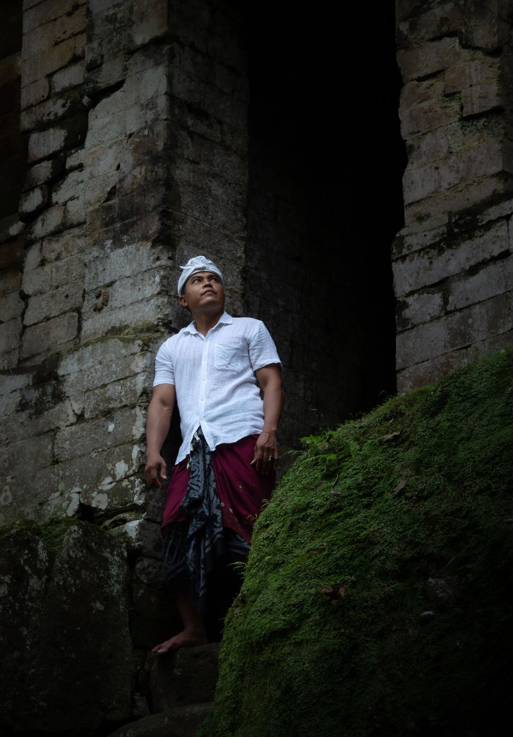 Figure in white shirt and headwrap stands at mossy stone ruins entrance.