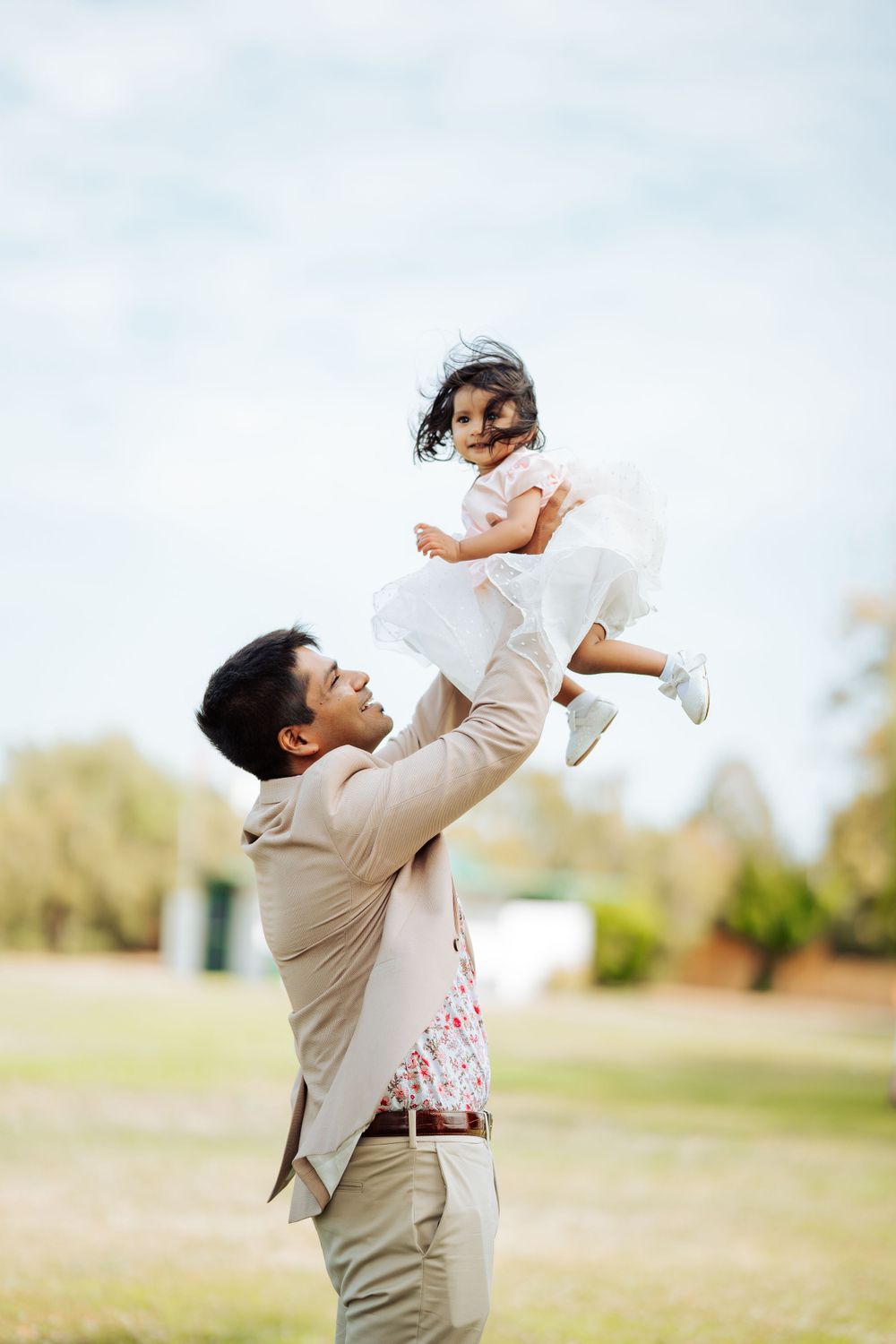 Parent lifting small child wearing white dress into the air at a park during golden hour.