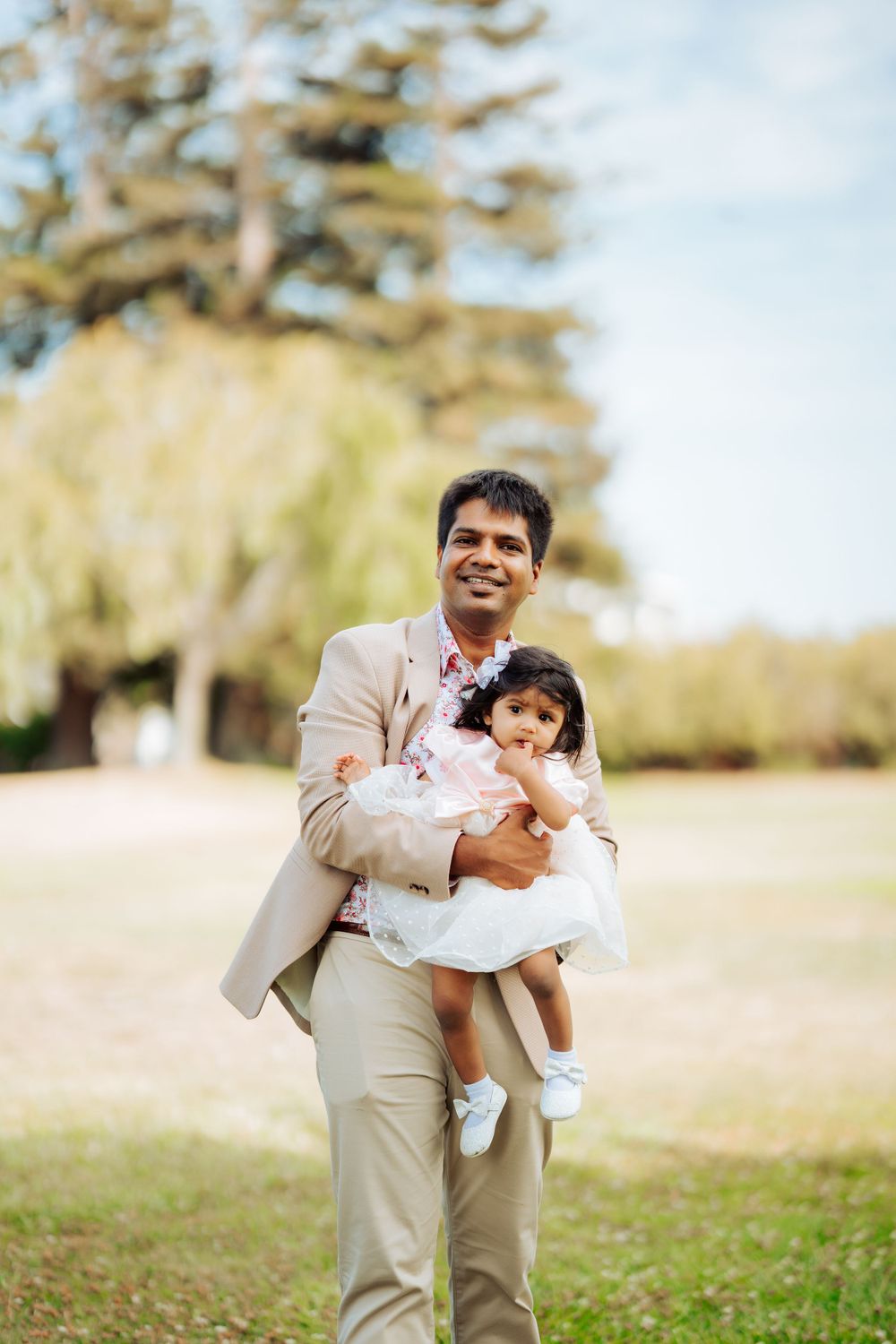 Parent in beige suit holding toddler in white dress outdoors in park setting.
