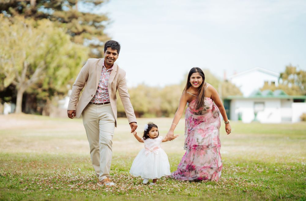 Family enjoying a playful moment together in a sunny park with parents holding hands with small child in white dress.