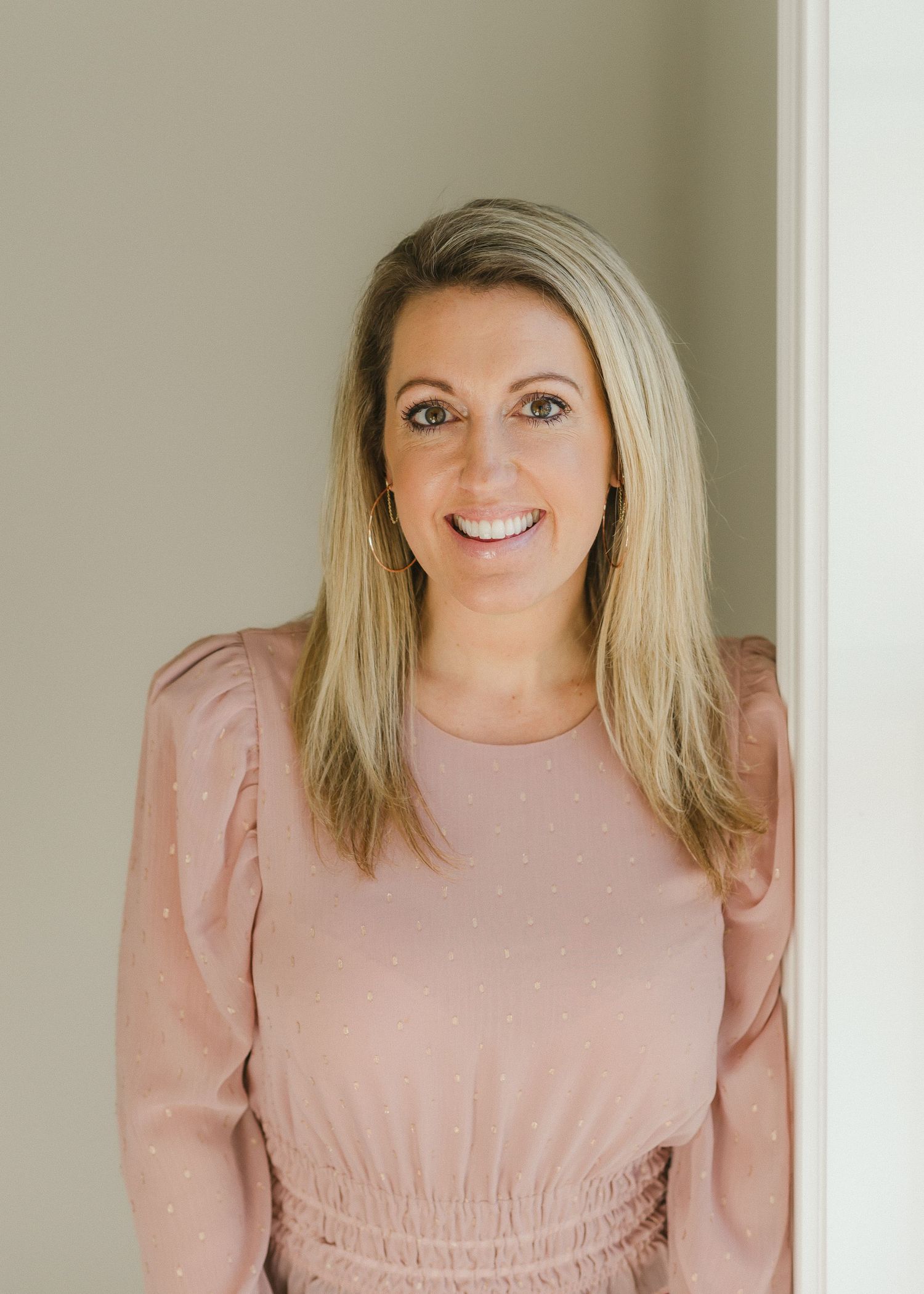Woman in pink blouse smiles warmly against neutral wall backdrop.