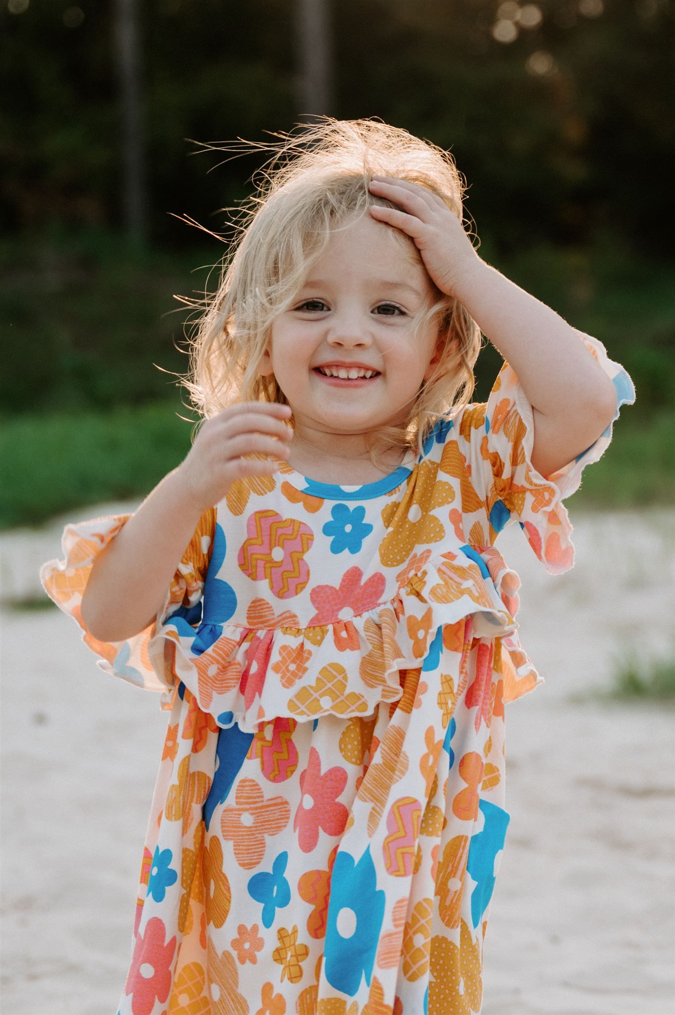 Young child in floral dress smiles and touches hair while standing on sandy ground.