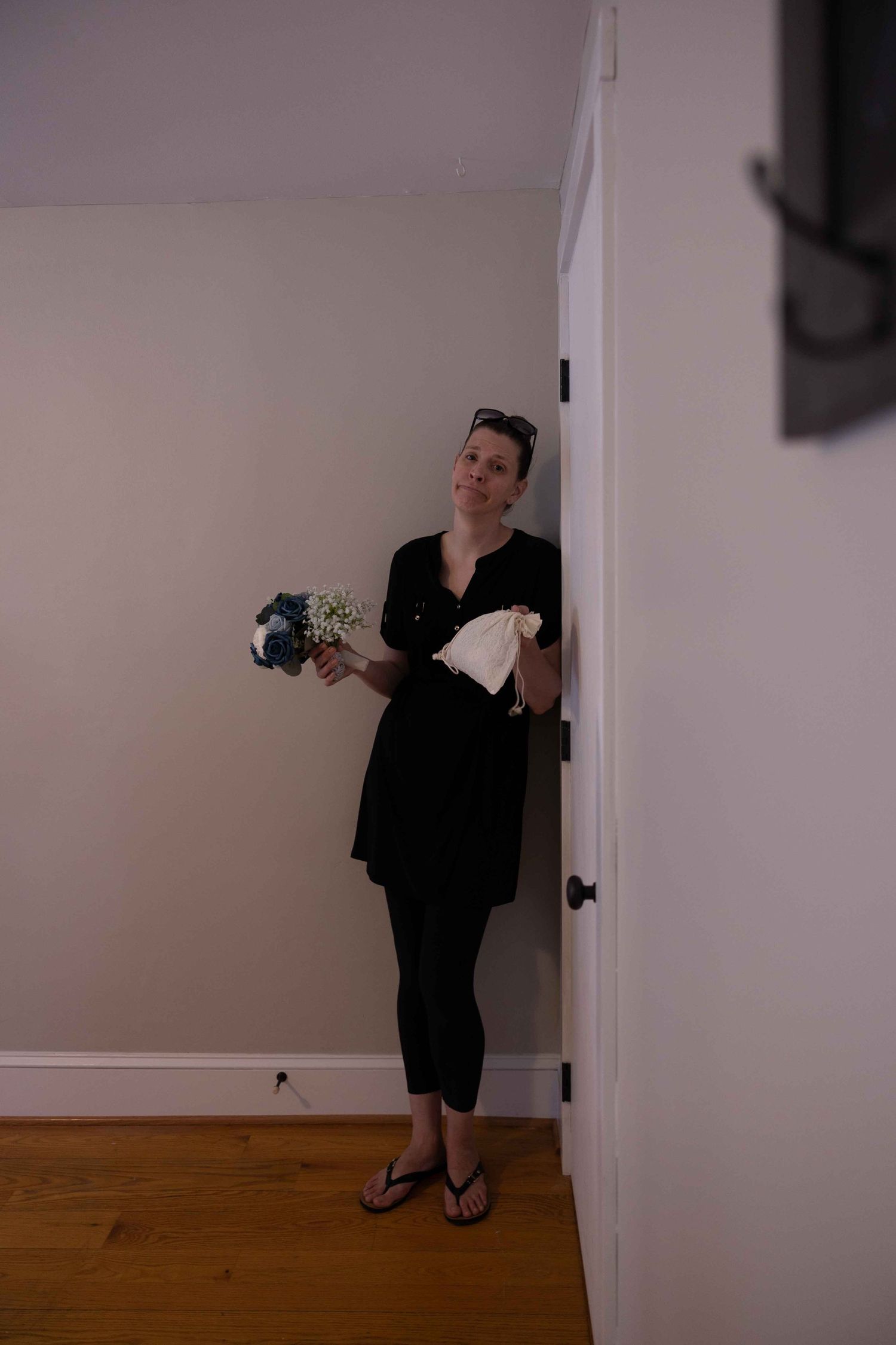 A person in black attire holds flowers while standing against a white wall.