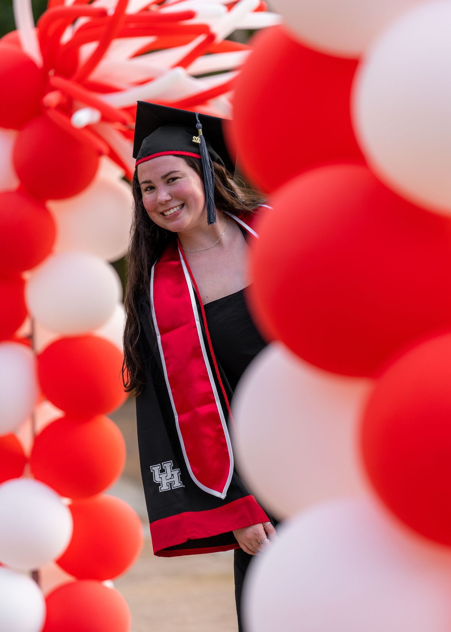 Graduate in cap and gown framed by red and white balloon arch during celebration.