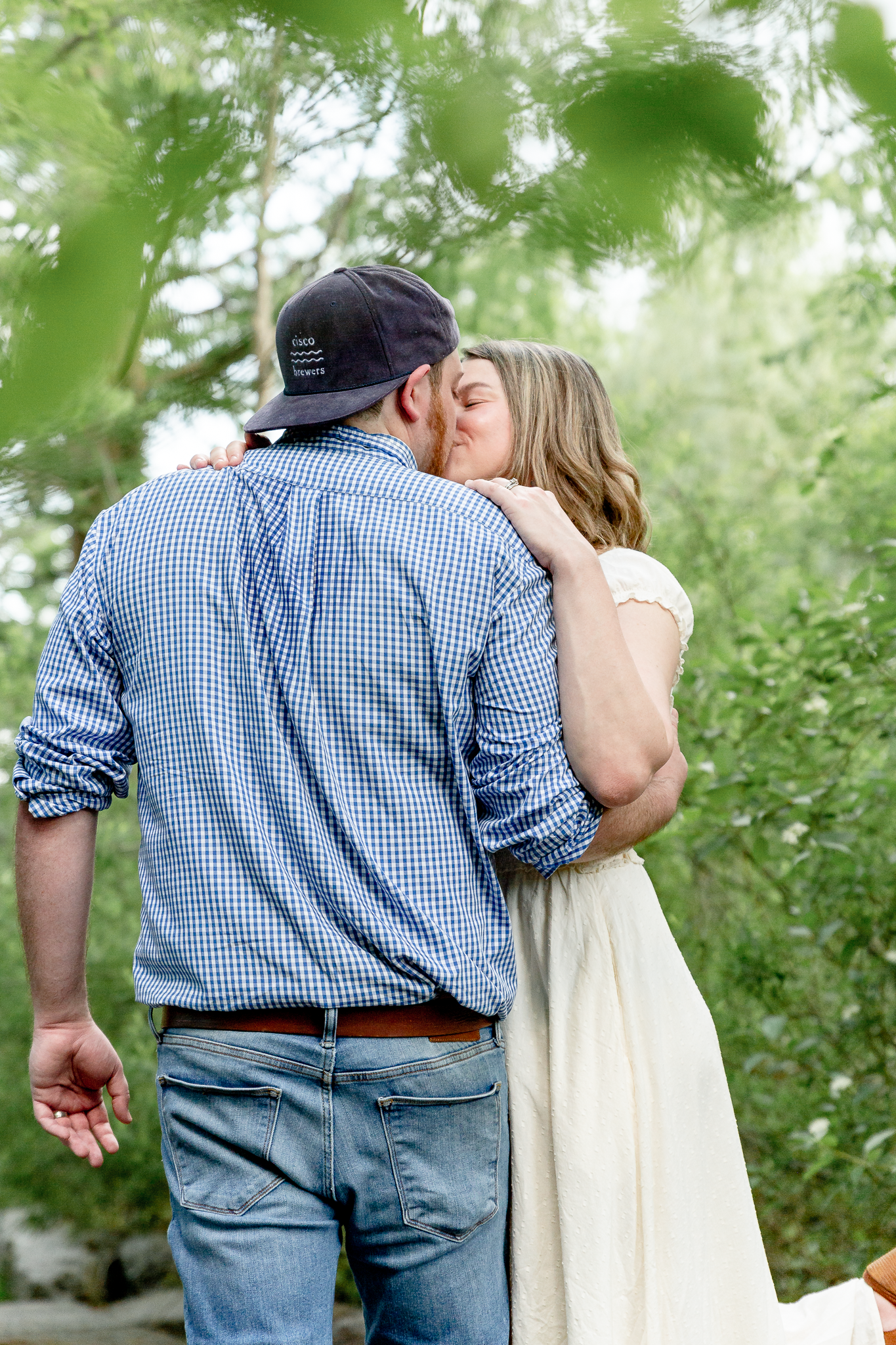 man in blue shirt leaning in for kiss to woman with white dress through trees with leg in the air,