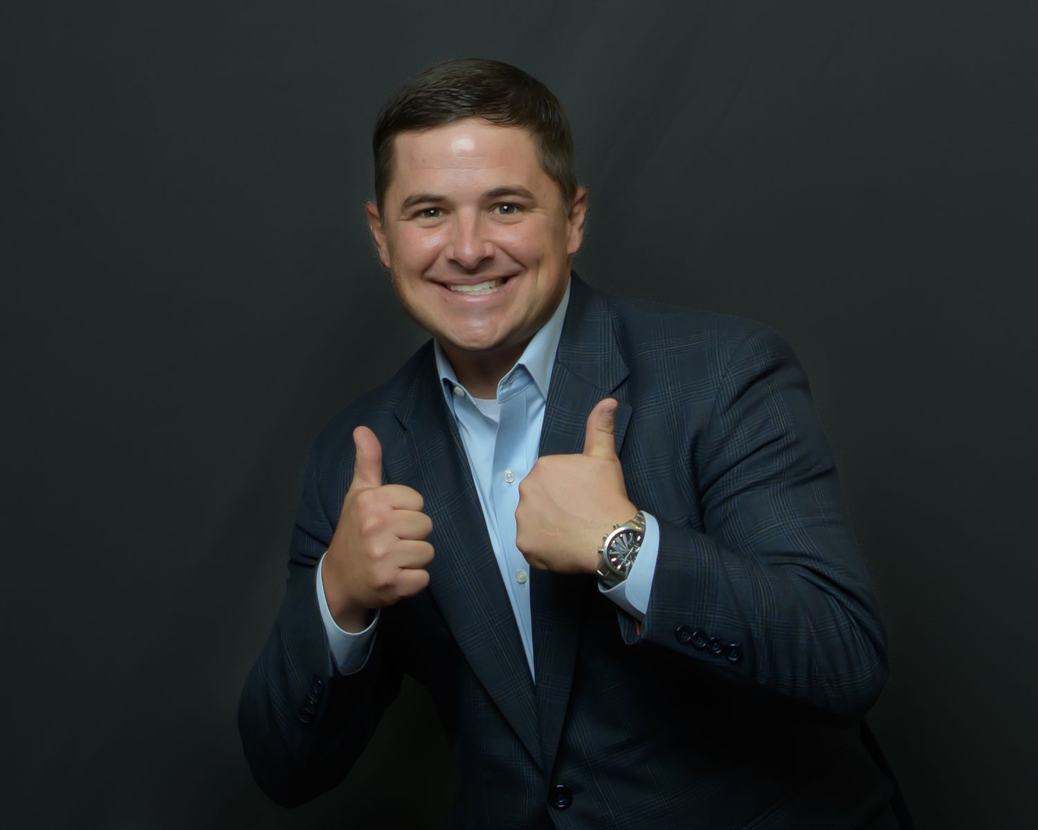 Professional portrait of person in navy suit giving thumbs up against dark background.