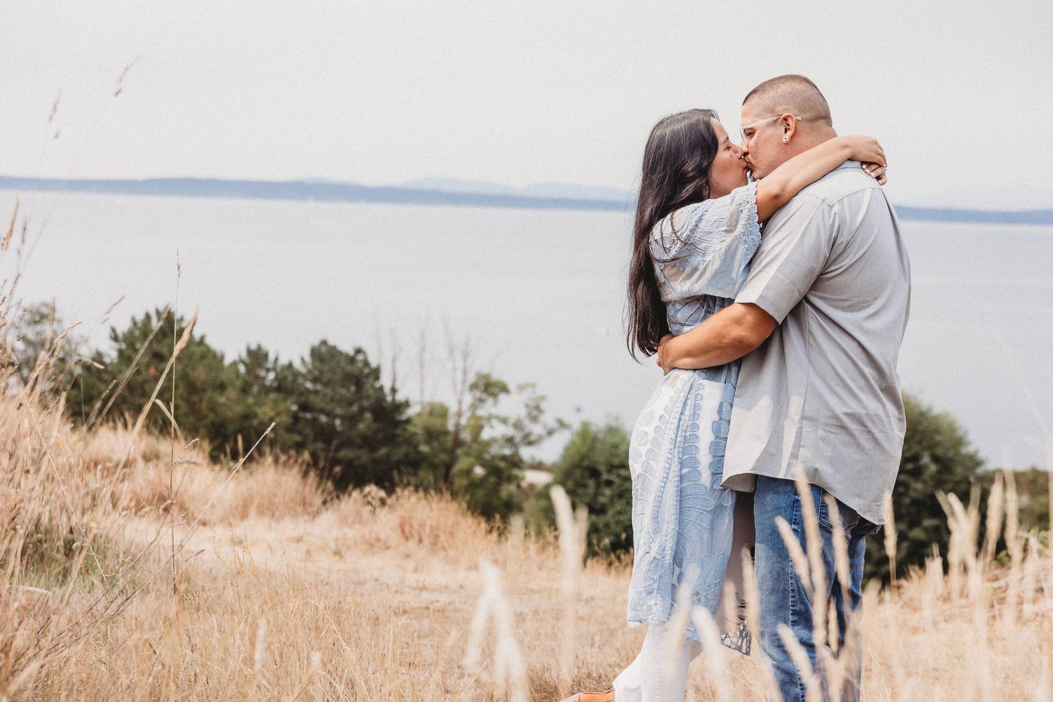 A couple shares an intimate moment in tall grass with a waterfront view at sunset.