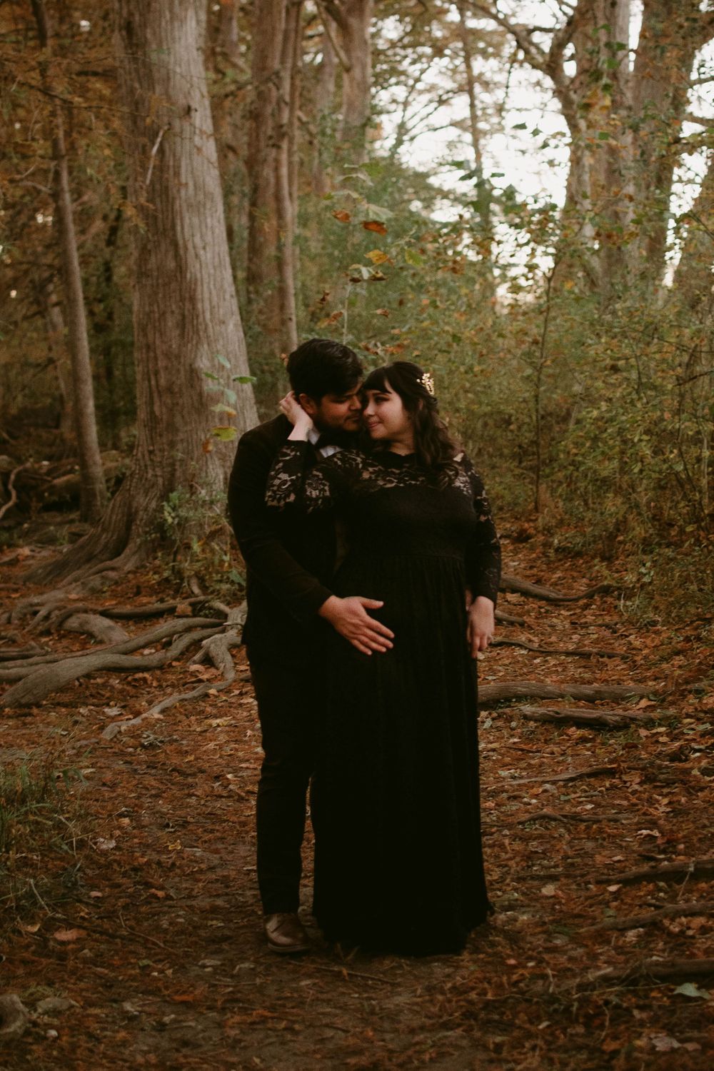 A romantic couple in black attire embraces tenderly among tall trees in a moody forest setting.