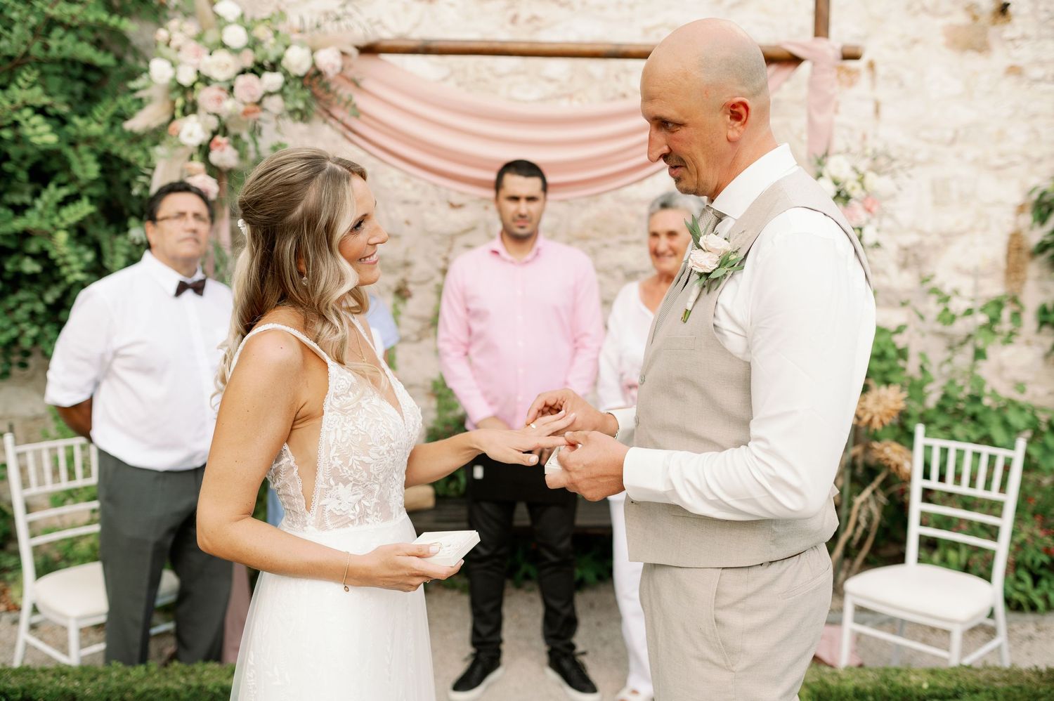 Intimate outdoor wedding ceremony with peach draping backdrop and white garden chairs in a stone courtyard setting.