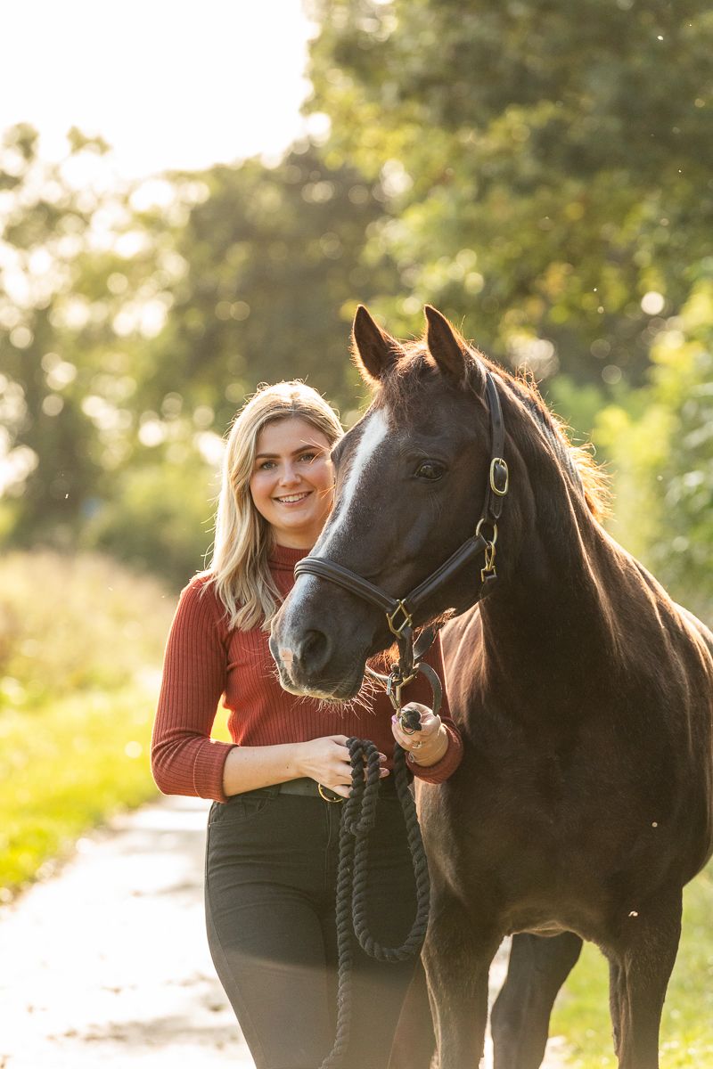 A dark horse and person pose together on a sunlit path during golden hour.